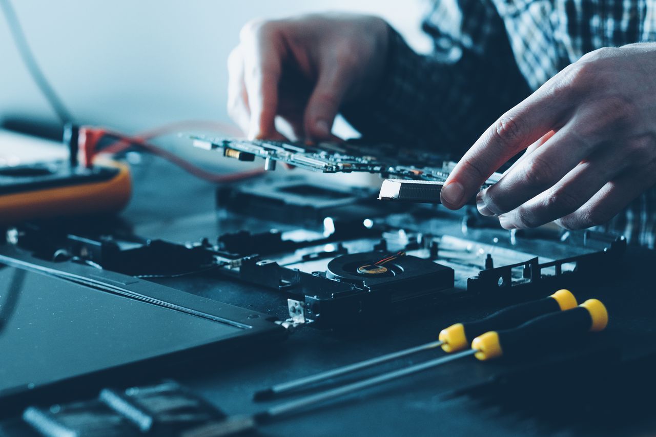 Zwei Hände heben eine Platine aus einem geöffneten Notebook, daneben Lüfter und Schraubendreher